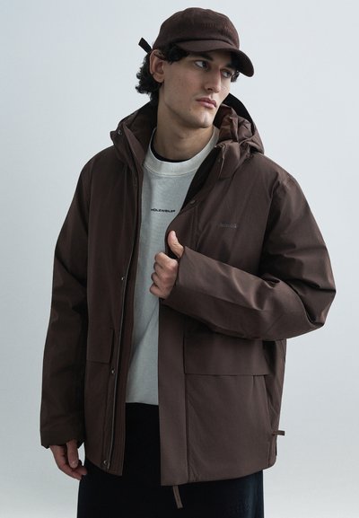 Veste marron imperméable avec capuche, dotée d'une fermeture éclair à l'avant, de deux grandes poches et d'un logo sur la poitrine. Portée par-dessus un pull léger.