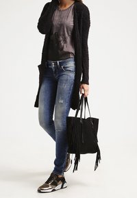 Black textured cardigan, gray graphic t-shirt, distressed blue jeans, black fringe tote bag, and silver sneakers with black accents.