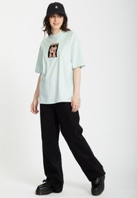 Hellblaues T-Shirt mit kurzen Ärmeln und grafischem Druck, kombiniert mit schwarzen weit geschnittenen Hosen und schwarzen Plateauschuhen. Lässiger Streetwear-Stil.