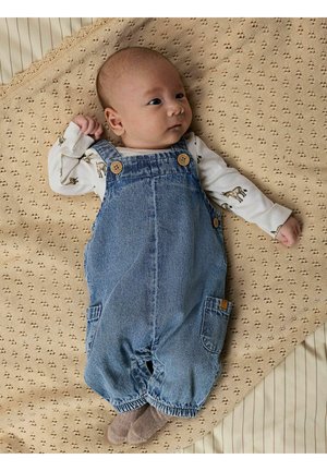 Peto vaquero con botones de madera y bolsillos laterales, usado sobre una camisa blanca de manga larga con estampados de animales; fondo beige suave con textura.