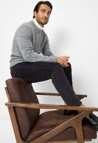 Grå strikket sweater med ribbede manchetter, mørke velourbukser og sorte sko, siddende på en stol med brun læderpolstring og træarmlæn.