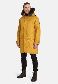 Parka isolata gialla con cappuccio foderato in pelliccia, tasche frontali, chiusura con zip e dettagli neri, abbinata a pantaloni neri e stivali stringati marroni.
