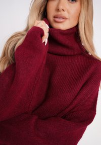 Burgundowa sweter z golfem o prążkowanej teksturze i oversize'owym kroju. Tkanina jest miękka i przytulna, z luźnym dopasowaniem w okolicy ramion i rąk.