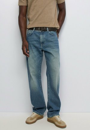 Homme portant un jean bleu ample, un t-shirt beige, une ceinture marron et des baskets beiges, debout devant un fond clair uni.