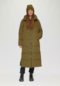 Lange olijfgroene pufferjack met hoge kraag, voorritssluiting en zijsplitten. Gecombineerd met een bijpassende gebreide beanie en zwarte broek.