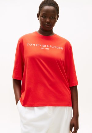 Donna di colore che indossa una T-shirt rossa Tommy Hilfiger con scritte bianche e pantaloni bianchi, in piedi davanti a uno sfondo bianco semplice.