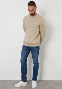 Hombre adulto con sudadera beige, jeans azules y zapatillas blancas, de pie con las manos detrás de la espalda contra una pared blanca sencilla.