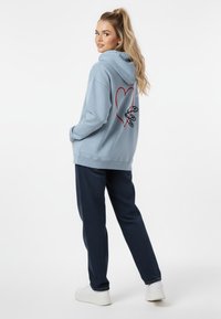 Felpa blu chiaro con design a cuore rosso e grafica sul retro; tessuto testurizzato. Abbinata a pantaloni blu navy scuro e scarpe da ginnastica bianche.