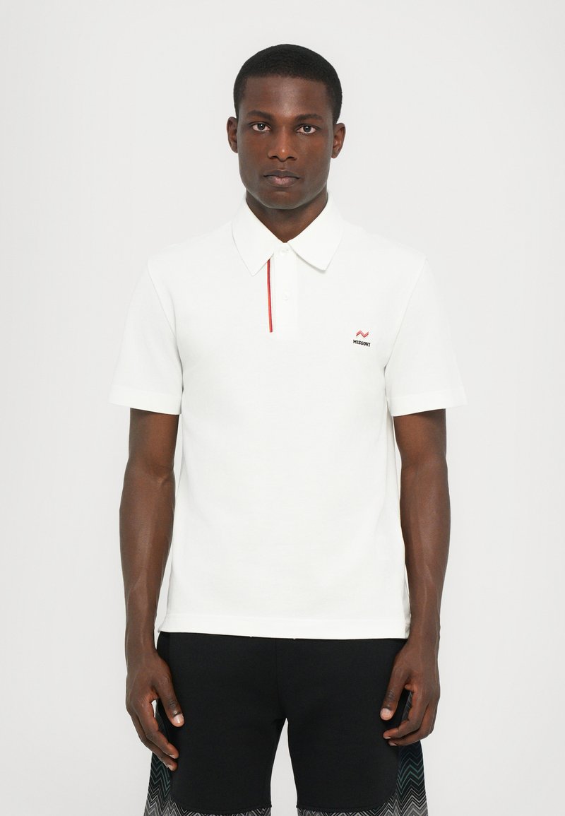 Camisa polo branca com um detalhe vermelho na pala, mangas curtas, tecido suave e um pequeno logotipo no lado esquerdo do peito. Design liso.