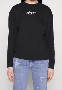 Černá mikina vyrobená z měkké textilie s kulatým výstřihem, spadlými rameny a bílým vyšívaným logem "Hugs" na hrudi. Vynesena s modrými roztrhanými džíny.