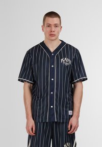 Camisa de estilo béisbol azul marino con rayas verticales blancas, escote en V, mangas cortas y cierre de botones. Presenta un logo en el pecho y una etiqueta en el dobladillo.