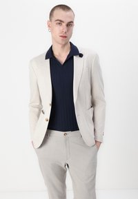 Lys beige blazer med to knapper og lommer, lagd over en marineblå ribbestrikket polo. Lyse grå bukser fullfører antrekket, med en knappdetalj.