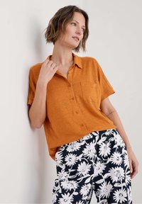 Korte mouwen, geknoopte blouse in oranje linnen met een borstzak, gecombineerd met zwarte broek met een wit bloemenpatroon.