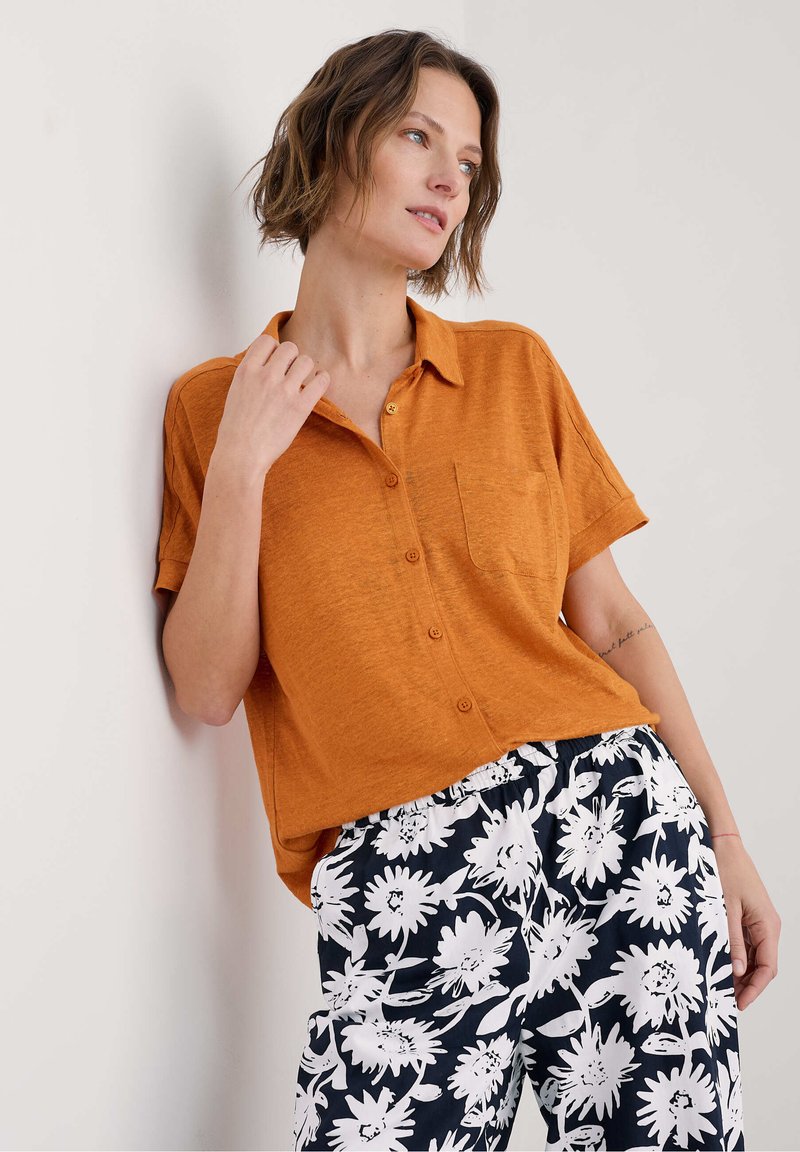Korte mouwen, geknoopte blouse in oranje linnen met een borstzak, gecombineerd met zwarte broek met een wit bloemenpatroon.