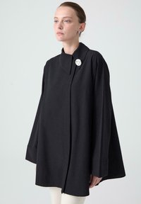 Black, loose-fitting outerwear with a high collar, long, flared sleeves, and a decorative silver button accent on the front.