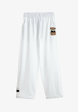 Joven con camiseta blanca de gran tamaño con estampado de oso de peluche, pantalones blancos a juego y zapatos marrones con suela gruesa, de pie sobre fondo blanco.