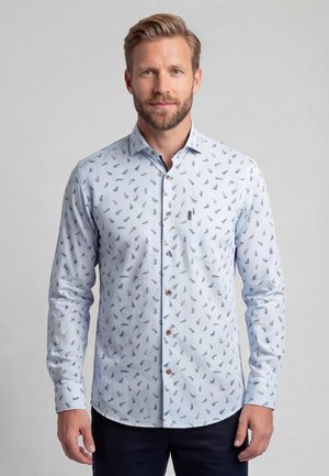 Man met kort blond haar en baard, draagt een lichtblauw overhemd met knopen met een patroon van kleine zeilboten tegen een effen achtergrond.