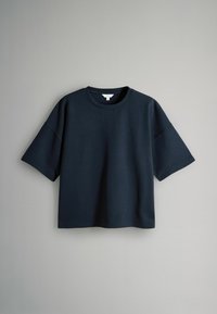 Tmavě námořnické oversized tričko s krátkým rukávem a kulatým výstřihem visící na pozadí jednoduché světle šedé zdi.