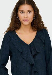 Navyblauwe blouse met een V-hals en gerimpelde accenten aan de voorkant. Gemaakt van textuurstof met subtiele strepen en lange mouwen.
