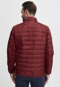 Giacca imbottita bordeaux con tessuto testurizzato sui lati, caratterizzata da un collo alto e sottili strisce bianche. Stile cropped, design leggero.