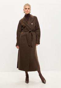 Manteau long marron avec une taille ceinturée, boutons double boutonnage, col cranté et poches latérales. Le tissu semble texturé et chaud.