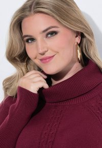 Pull en maille bourgogne avec un col montant et des détails en câble texturés, featuring des manchons côtelés. Accessoirisé avec de grandes boucles d'oreilles en or.