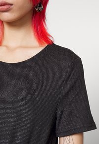 Personne aux cheveux rouge vif portant un top noir texturé à manches courtes, une grande boucle d'oreille florale noire et un tatouage en ligne visible sur l'avant-bras.