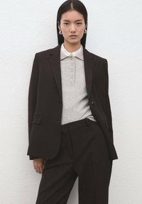 Brązowa, dopasowana marynarka noszona na szarej koszulce polo z guzikami, zestawiona z pasującymi brązowymi spodniami. Gładka tkanina o minimalistycznym designie.