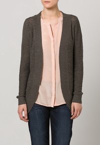 Grau gestrickte Cardigan mit geripptem Muster und offener Vorderseite, getragen über einer hellrosa Bluse. Enganliegende Ärmel und lässiger Schnitt. Denim-Jeans sind sichtbar.