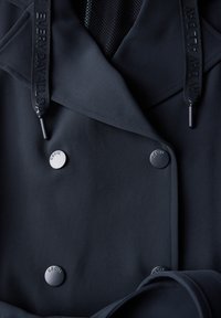 Černý trench coat s hladkým materiálem. Obsahuje dvojité zapínání na knoflíky, šňůrky s logem a elegantní design. Viditelné texturované podšívka.