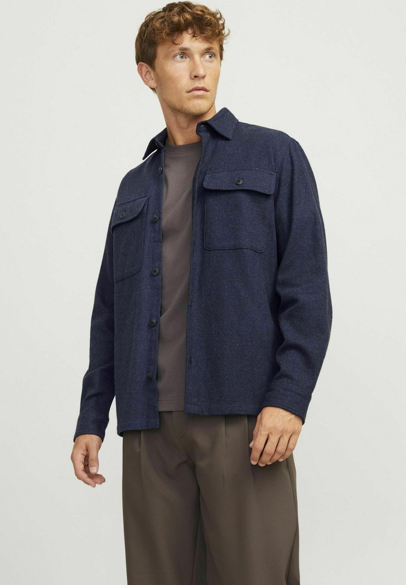 Giacca camicia in misto lana blu navy con chiusura a bottoni, due tasche sul petto e colletto. Il modello indossa una T-shirt grigia e pantaloni marroni.
