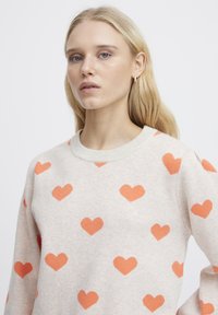 Ljusbeige tröja med ett mönster av orangea hjärtan. Tillverkad av ett mjukt stickat material med ribbad halsringning och avslappnad passform.