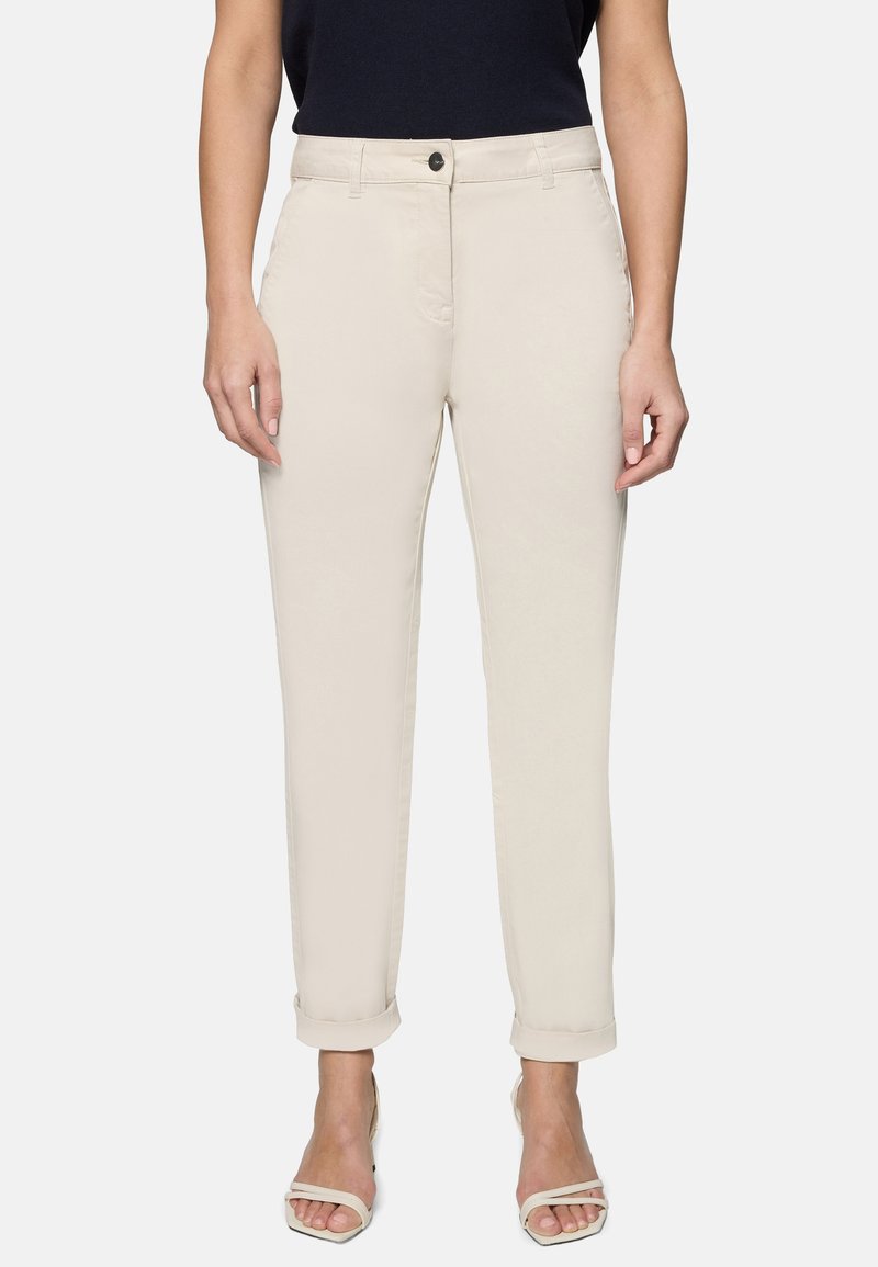 Femme portant un pantalon beige droit avec revers, un haut noir et des sandales à talons ouvertes beiges, debout devant un fond blanc.