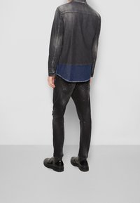 Giacca di jeans in nero sbiadito con parte inferiore in denim blu, abbinata a jeans neri strappati e stivali neri. Cuciture e hardware visibili.