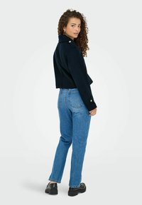 Marineblå beskåret jakke med fremtrædende skulderknapper, parret med højformat blå jeans og sorte klodsede sko, der fremviser et afslappet look.