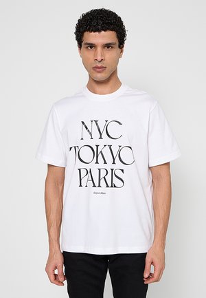 Uomo con capelli ricci che indossa una maglietta bianca con la scritta "NYC TOKYO PARIS" e pantaloni neri, in piedi davanti a uno sfondo grigio chiaro.