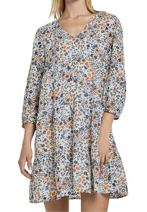 Kobieta w sukience do kolan z dekoltem w szpic i rękawami do 3/4, ozdobionej małym, wielokolorowym wzorem kwiatowym w odcieniach niebieskiego, pomarańczowego, czarnego i jasnego żółtego.