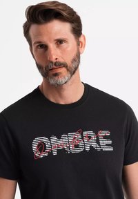 Sort kortærmet t-shirt med "OMBRE" trykt i hvid og rød, med et stribet design i fed skrift. Blødt stof og rund hals.