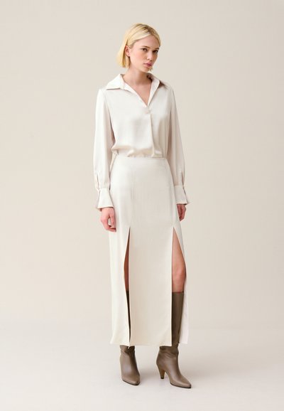 Blusa di raso crema con colletto a punta, abbinata a una gonna maxi coordinata con due spacco alta coscia. Stivali con tacco marroni fino al ginocchio.