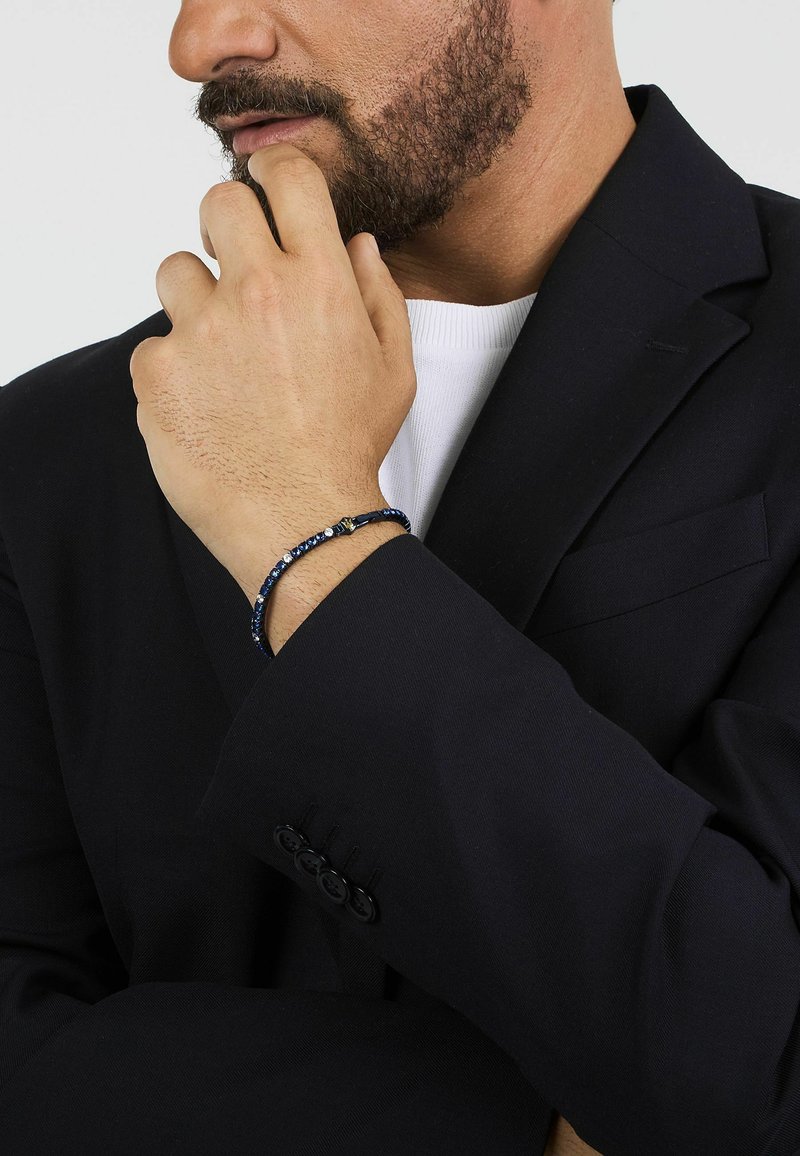 Marinblå pärlarmband med små silverdetaljer, bärs på handleden, ihop med en svart kavaj och vit skjorta. Slät textur och enkel design.