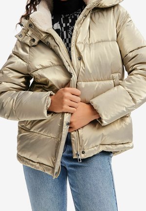 Person trägt metallisch beige gesteppte Jacke mit Lammfellkragen, die er zumacht, kombiniert mit blauen Jeans und einem schwarzen gemusterten Oberteil.