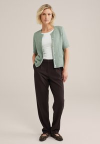 Cardigan texturé vert menthe à manches courtes, fermeture à boutons, porté sur un top blanc et associé à un pantalon noir et des ballerines accentuées.