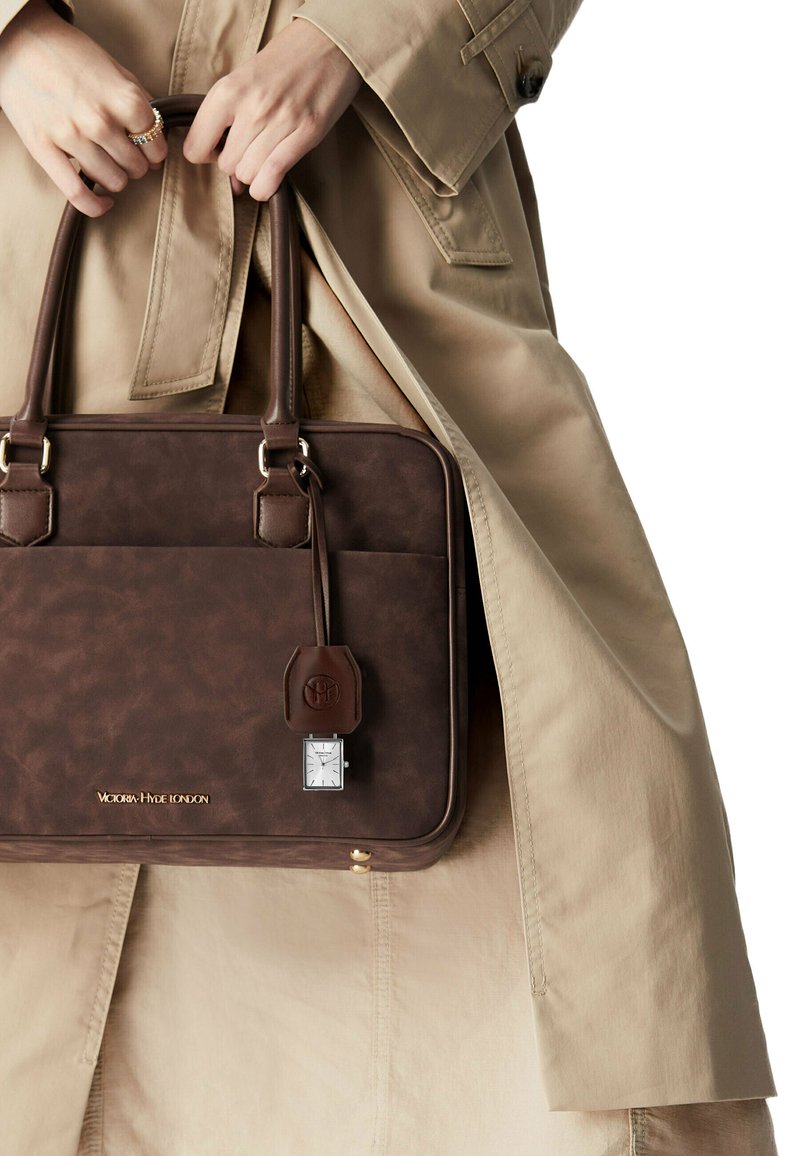 Braune Lederhandtasche mit goldenen Beschlägen, rechteckiger Form, Fronttasche und einem abnehmbaren Schlüsselanhänger in Form einer silbernen Uhr. Eine beige Trenchcoat ist sichtbar.