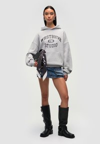 Felpa grigia con stampa "WRSTBHVR STUDIO", mini gonna di jeans, stivali neri fino al ginocchio con fibbie e una sciarpa nera con motivi bianchi.