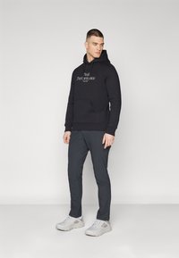 Sudadera negra con bolsillo frontal y logotipo bordado, combinada con pantalones gris oscuro y zapatillas gris claro, todo sobre un fondo liso.