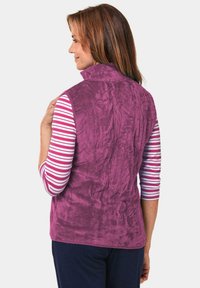 Fleece-Weste in Lila mit hohem Kragen und strukturierter Oberfläche. Über einem gestreiften Langarmshirt getragen. Die Rückansicht zeigt das nahtlose Design.