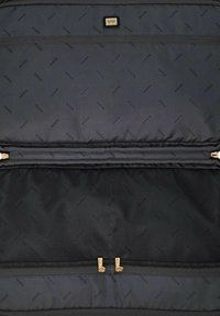 Sac en tissu noir avec garnitures dorées, comportant un compartiment zippé et un motif logo. L'intérieur est doublé de mesh avec une section zippée supplémentaire.