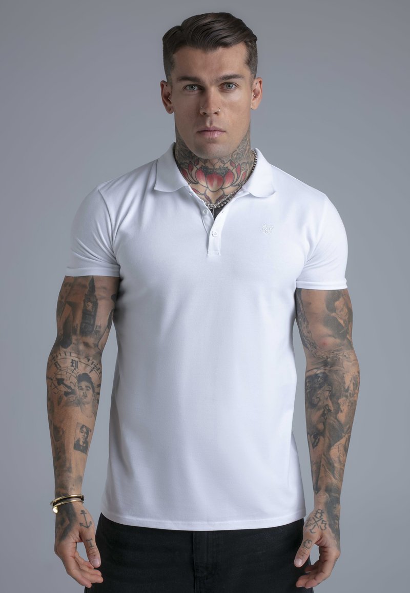 Hombre con tatuaje floral en el cuello y mangas completas de retratos, vistiendo una camisa polo blanca y pantalones negros, de pie contra un fondo gris liso.