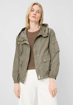 Blonde Frau trägt einen braunen Strickpullover, eine khakigrüne Kapuzenjacke mit Taschen und weiße Hosen, steht vor einem schlichten grauen Hintergrund.