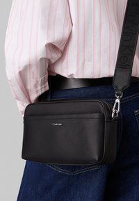Juodas odinis per petį nešiojamas krepšys su lygiu paviršiumi, sidabro spalvos detalėmis ir nuimama petneša. Priekyje yra subtilus Calvin Klein logotipas.